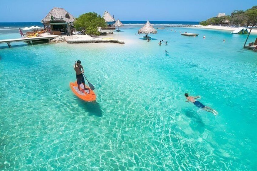 A person paddleboarding in clear turquoise water, another person swimming, and a beach with thatched-roof huts and boats in the background. 3
