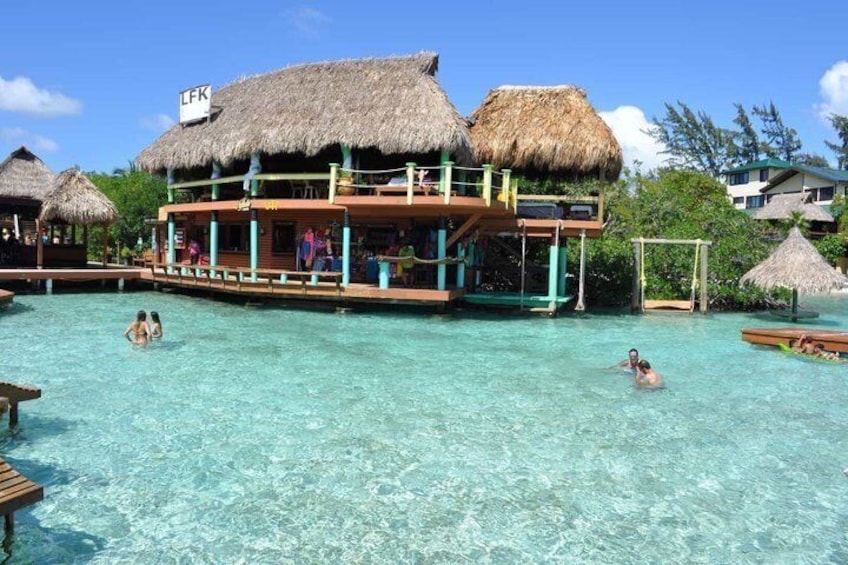 A tropical resort with a clear pool, thatched-roof structures, and a wooden building with a sign. 4