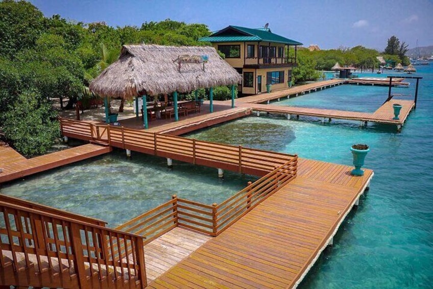 A tropical resort with wooden walkways over clear blue water, a thatched-roof building, and lush greenery. 1