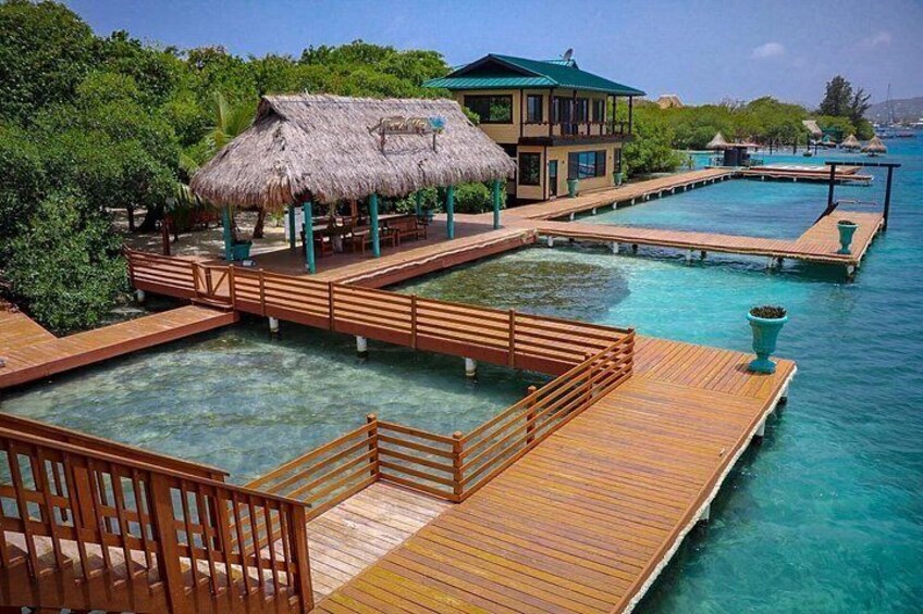 A tropical resort with wooden walkways over clear blue water, a thatched-roof building, and lush greenery. 1