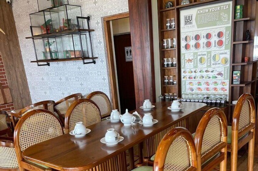 A wooden dining table set with white teapots and cups, surrounded by wicker chairs. A glass display case with various items is mounted on the wall, and a 'Know Your Tea' poster is visible in the background. 3