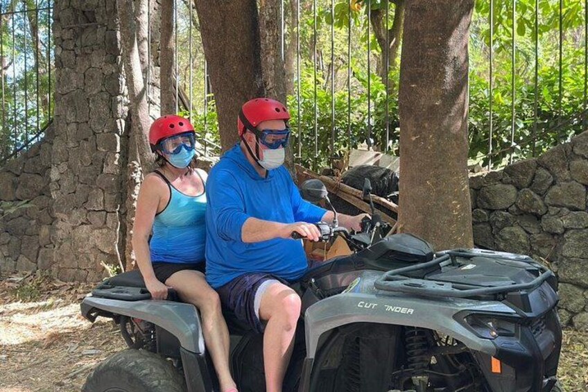 Two people on a quad bike in a forested area with trees and a stone wall. 5