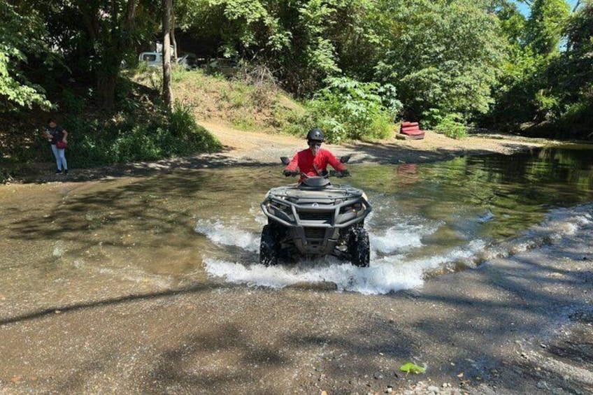 A person riding an ATV through shallow water on a road surrounded by trees. 3