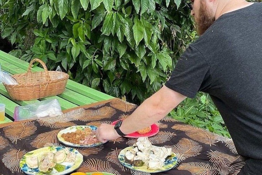 Una persona sentada a una mesa con platos de comida, una cesta y un fondo de follaje verde. 3