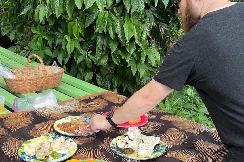 A person sitting at an outdoor table with plates of food, a basket, and a green leafy background. 3