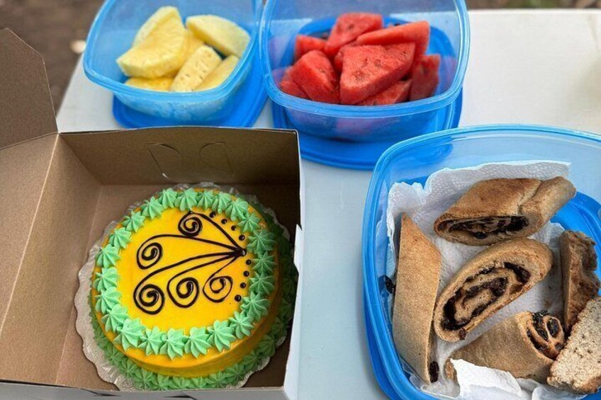 Un pastel con cobertura verde y base amarilla, una caja con un pastel, y recipientes con frutas y pasteles en porciones. 3
