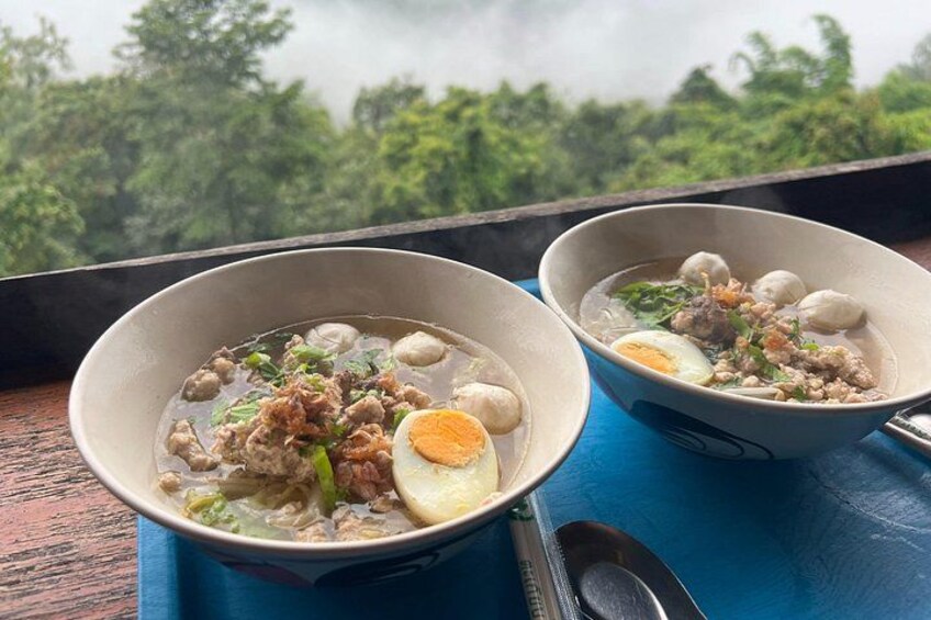 Two bowls of soup with meat and egg on a blue table with a view of trees. 2