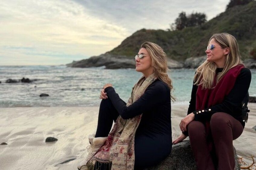 Twee personen zitten op een rots aan zee, met een glooiend landschap op de achtergrond. 3