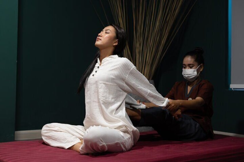 A person in white performing a seated yoga pose on a red mat, with another person assisting. 2