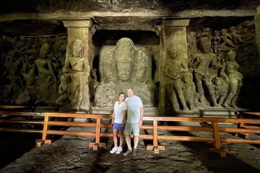A couple standing in front of ancient stone carvings, with a wooden railing in the foreground. 3
