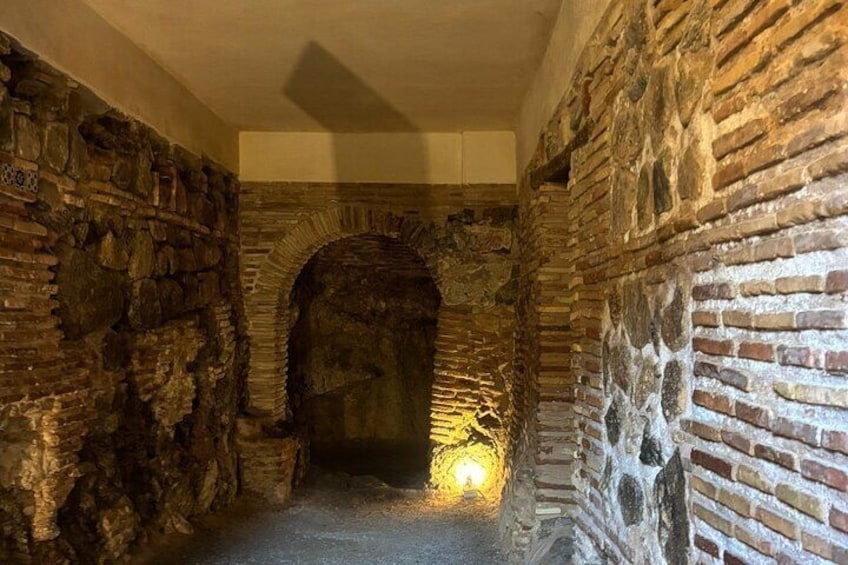 A stone corridor with an arched doorway and a light at the end. 1