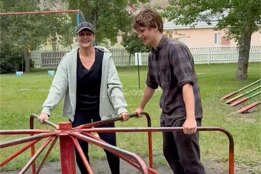 Two people pushing a red and black handcart in a grassy area with trees and a fence in the background. 4