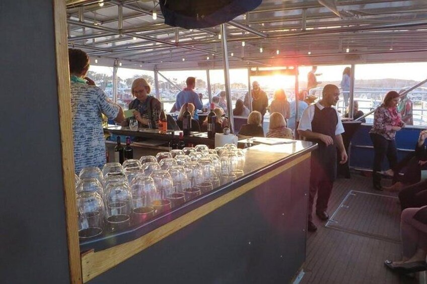A bar area on a boat with people seated and standing, a bartender serving drinks, and a row of empty glasses on the counter. 3