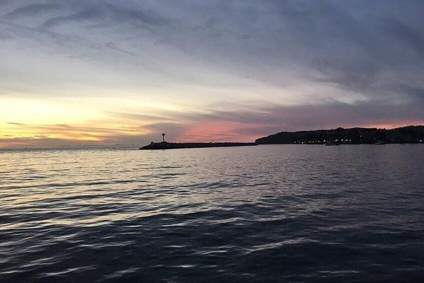 A calm sea at sunset with a lighthouse on a small island and a distant shoreline with buildings. 4