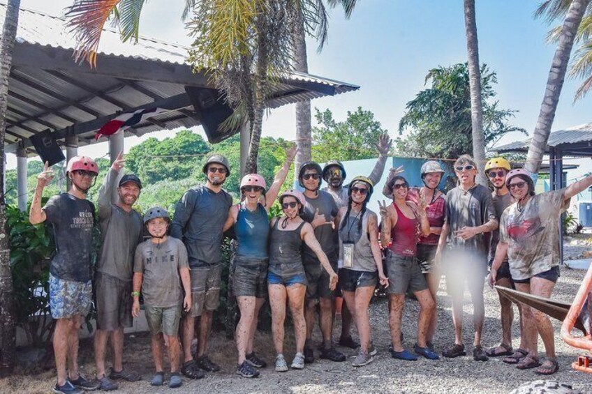 A group of people outdoors, some wearing helmets, standing under a shelter with palm trees in the background. 2