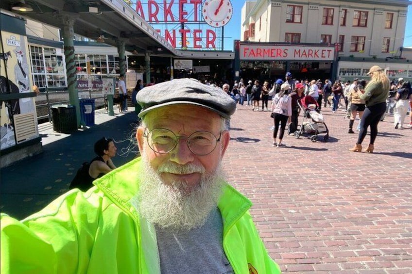 En livlig utomhusmarknad med ett klocktorn, en skylt som visar att det är en bondemarknad, och folk som promenerar runt. 1