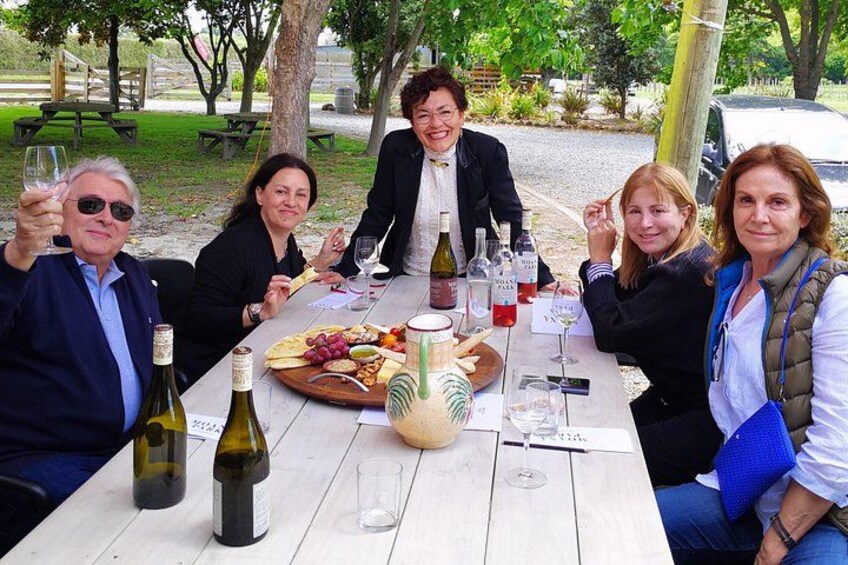 A group of people sitting at an outdoor table with wine bottles, glasses, and a cheese board. 2