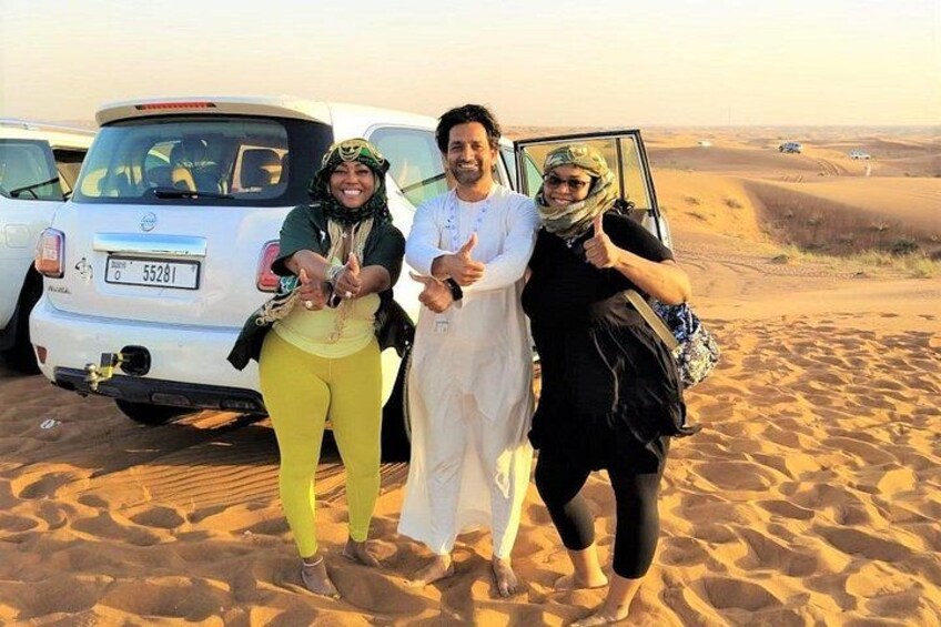 Three people in traditional attire standing on sand dunes with a white vehicle in the background. 3
