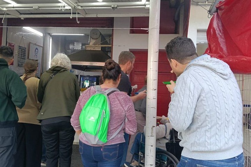 People standing in a public indoor space, possibly a waiting area or lobby, with a red wall and a white pillar. 2