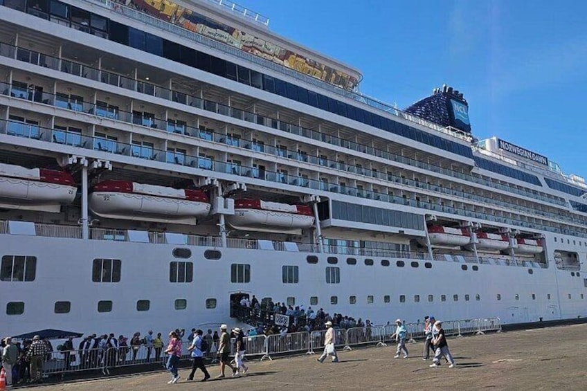A large cruise ship docked at a port with multiple decks and passengers visible on the lower levels. 1