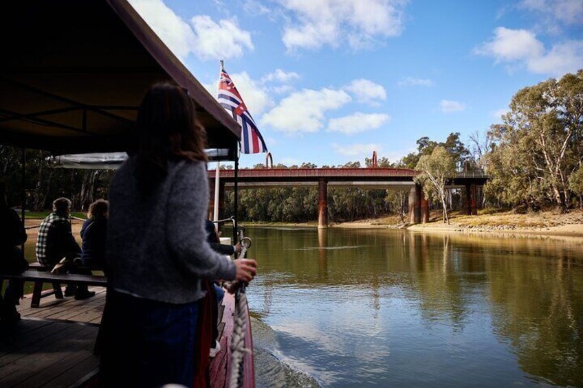 A person standing on a wooden deck overlooking a river, with a bridge and trees in the background. 4