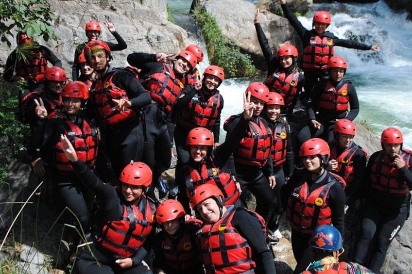 A group of people wearing red helmets and life vests, standing near a river with rocks and greenery. 2
