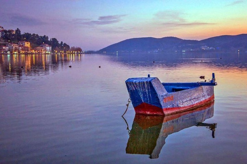 A calm lake with a small boat anchored near the shore, surrounded by buildings and hills in the background. 1