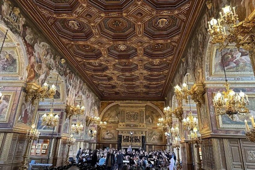 An ornate hall with a hexagonal patterned ceiling, chandeliers, and a crowd of people. 4