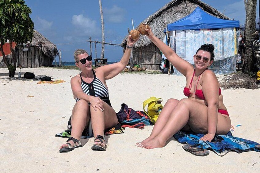 Two people sitting on a sandy beach with a thatched-roof hut and a blue tent in the background. 4