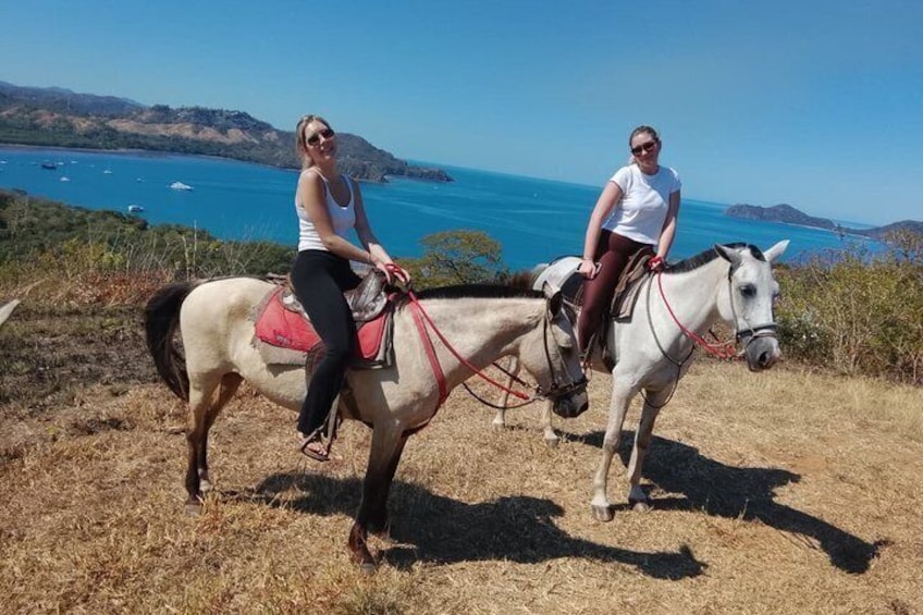 Two people riding horses on a hillside with a coastal view in the background. 5