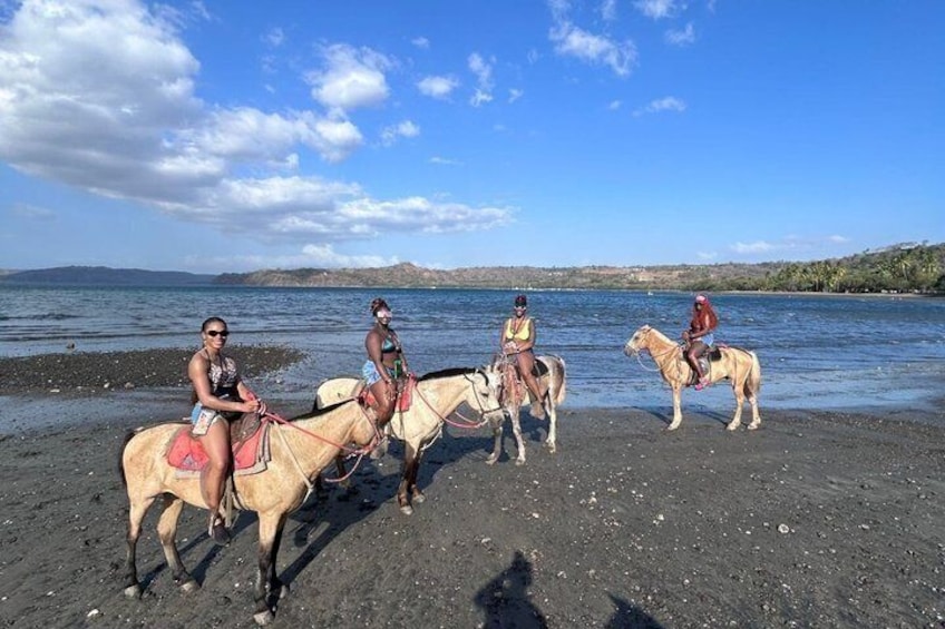 Four people riding horses on a beach with water in the background. 4