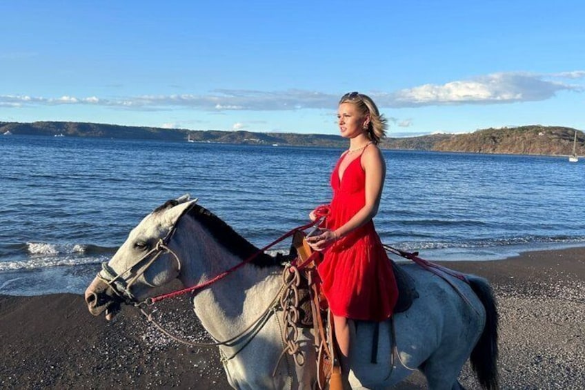A person in a red dress riding a white horse on a beach with water and hills in the background. 3