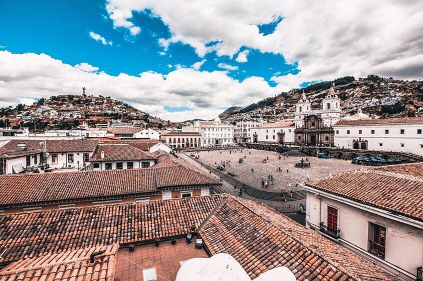 A cityscape with terracotta-roofed buildings, a large open square, and a hill with buildings. 5