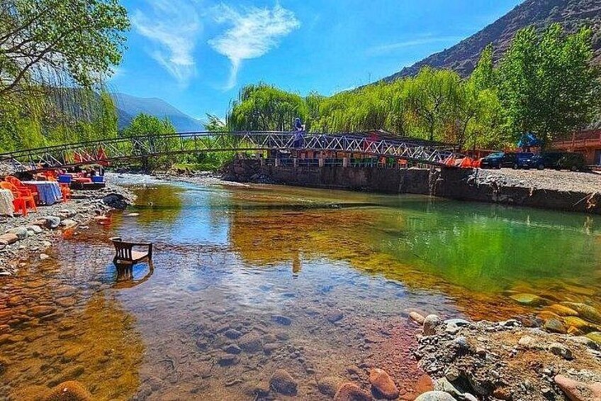 A clear river with rocks, a wooden chair, a bridge, and a building with a red roof. 1