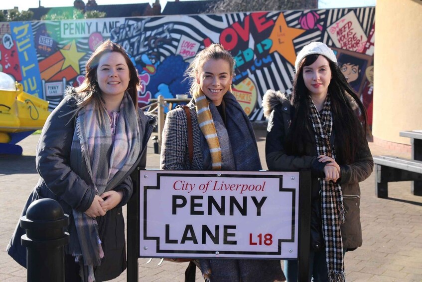Three people standing in front of a street sign for Penny Lane in Liverpool. 2