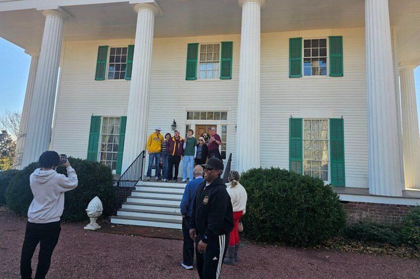 A historic building with white columns and green shutters, a group of people gathered on the steps, and a person taking a photo. 5