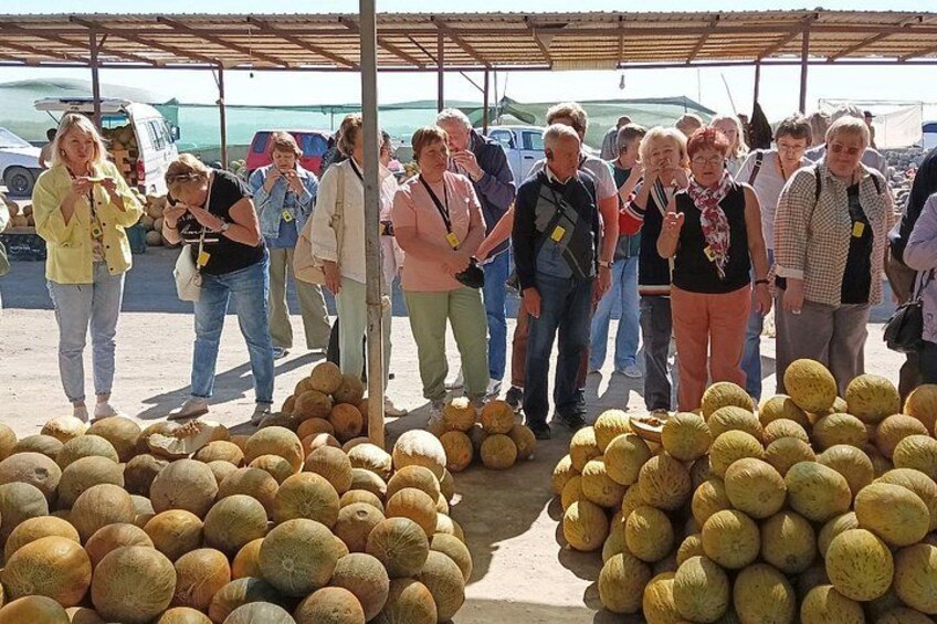 A group of people standing at a fruit market with large piles of melons. 1