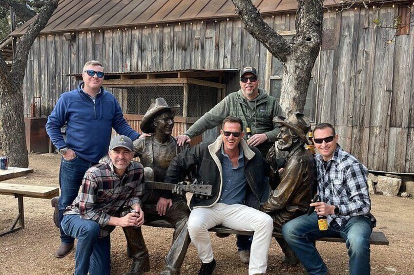 A group of people posing with a cowboy statue in an outdoor setting with a wooden building in the background. 3