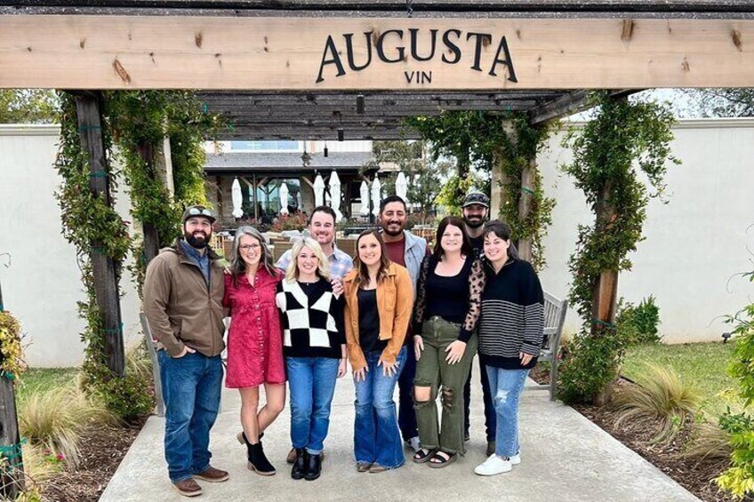 A group of people standing under a wooden archway with 'AUGUSTA VIN' written on it. 1