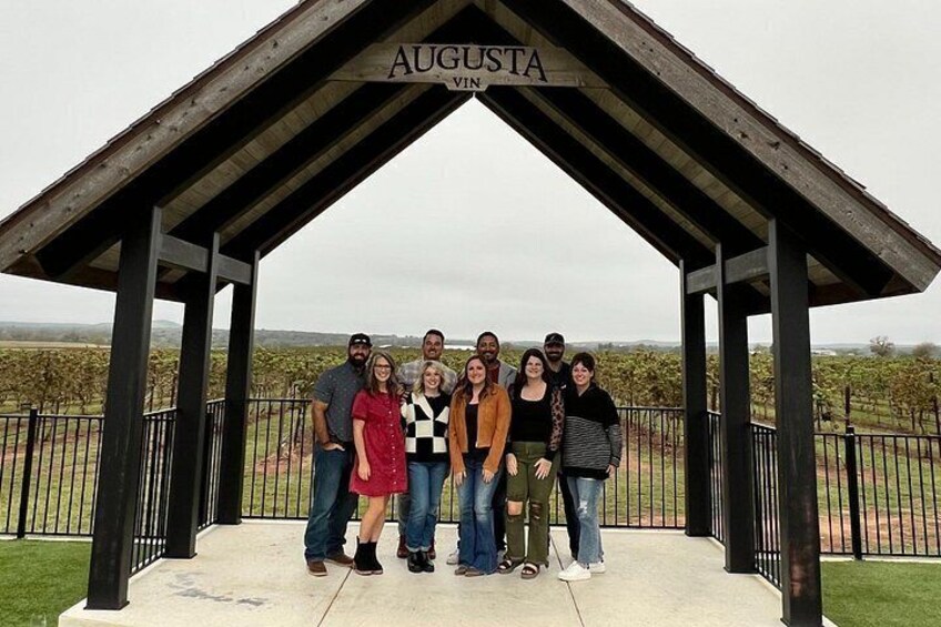 A group of people standing under a pavilion labeled 'AUGUSTA' with a vineyard in the background. 5