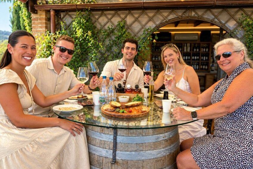A group of people sitting around a glass table outdoors with wine glasses, bottles, and plates of food. 1