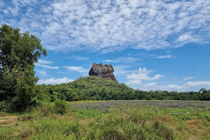 Une colline rocheuse surmontée d’une formation rocheuse imposante, entourée d’une végétation verdoyante et dense, et surplombée d’un ciel bleu clair parsemé de quelques nuages. 1