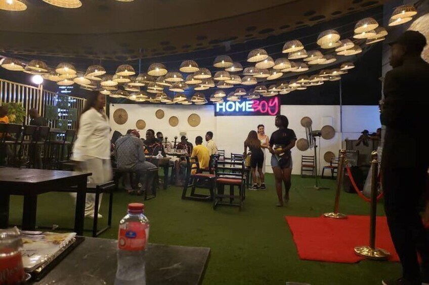 A social event with people mingling, a neon sign reading "HOMEBOY", and tables with drinks. 1