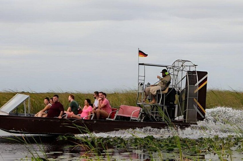 A boat with passengers and a flag, moving through water with vegetation. 2