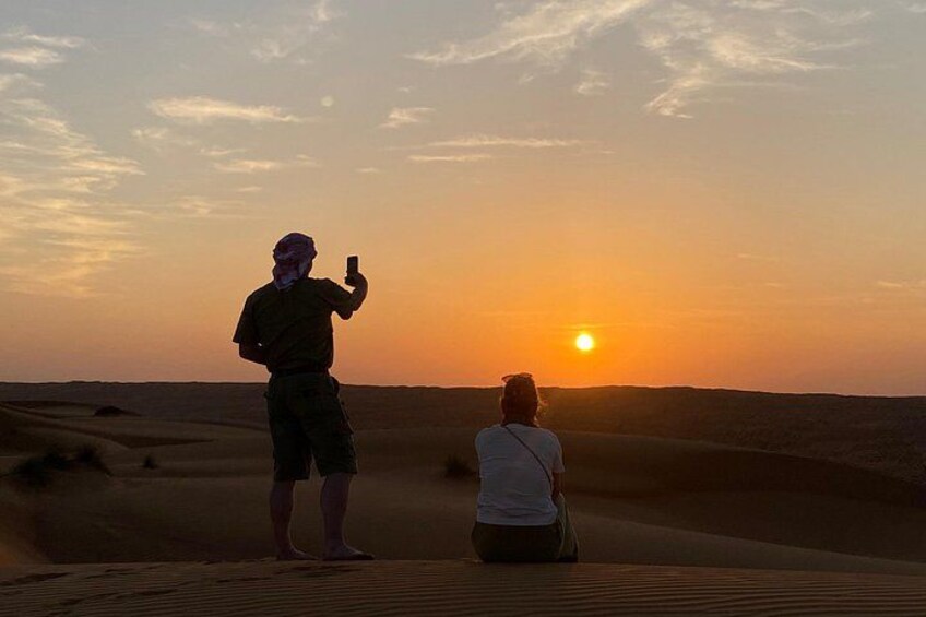 Due persone in piedi su una duna di sabbia, una delle quali sta scattando una foto, con il sole che tramonta sullo sfondo. 1