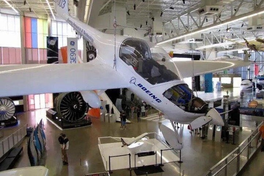 Een wit vliegtuig met de tekst 'BOEING' op de zijkant, hangend in een hangar met andere vliegtuigen en tentoonstellingen. 3