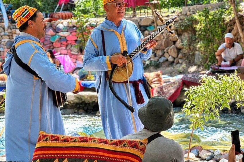 Deux personnes, vêtues de costumes traditionnels, jouent d’instruments à cordes, avec une rivière et des bateaux en arrière-plan. 5