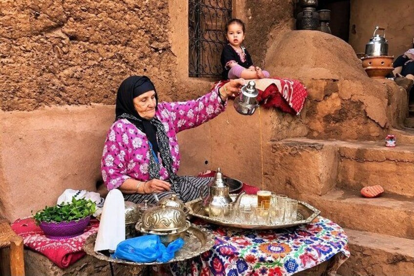 Une femme âgée, vêtue d’un costume traditionnel, sert du thé à un enfant dans un cadre rustique, avec des murs en terre et un pot en argile. 2