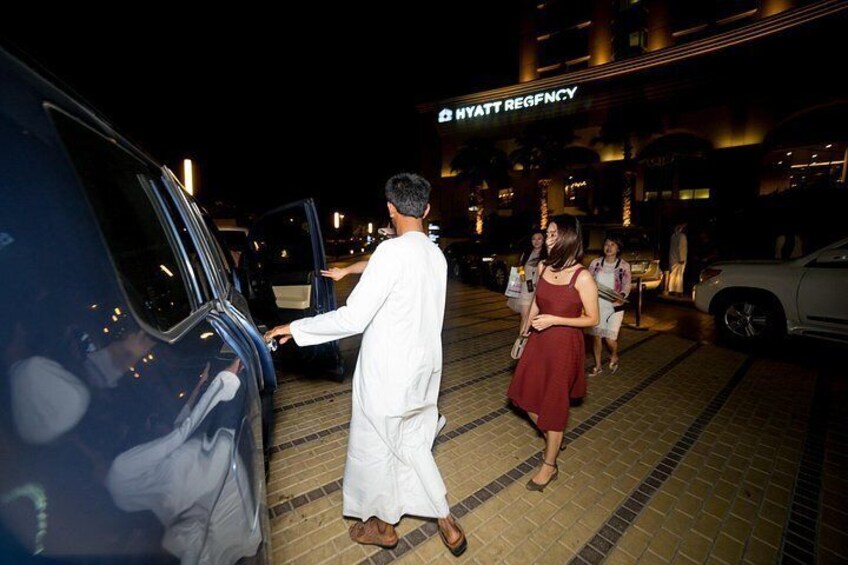 Un hombre con vestimenta tradicional sale de un vehículo azul, una mujer con un vestido rojo, y un edificio con un letrero que dice 'HYATT REGENCY' al fondo. 1