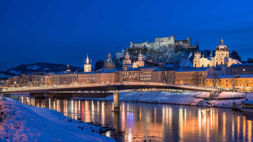 Un río cubierto de nieve con un puente, un castillo y edificios iluminados al fondo. 3